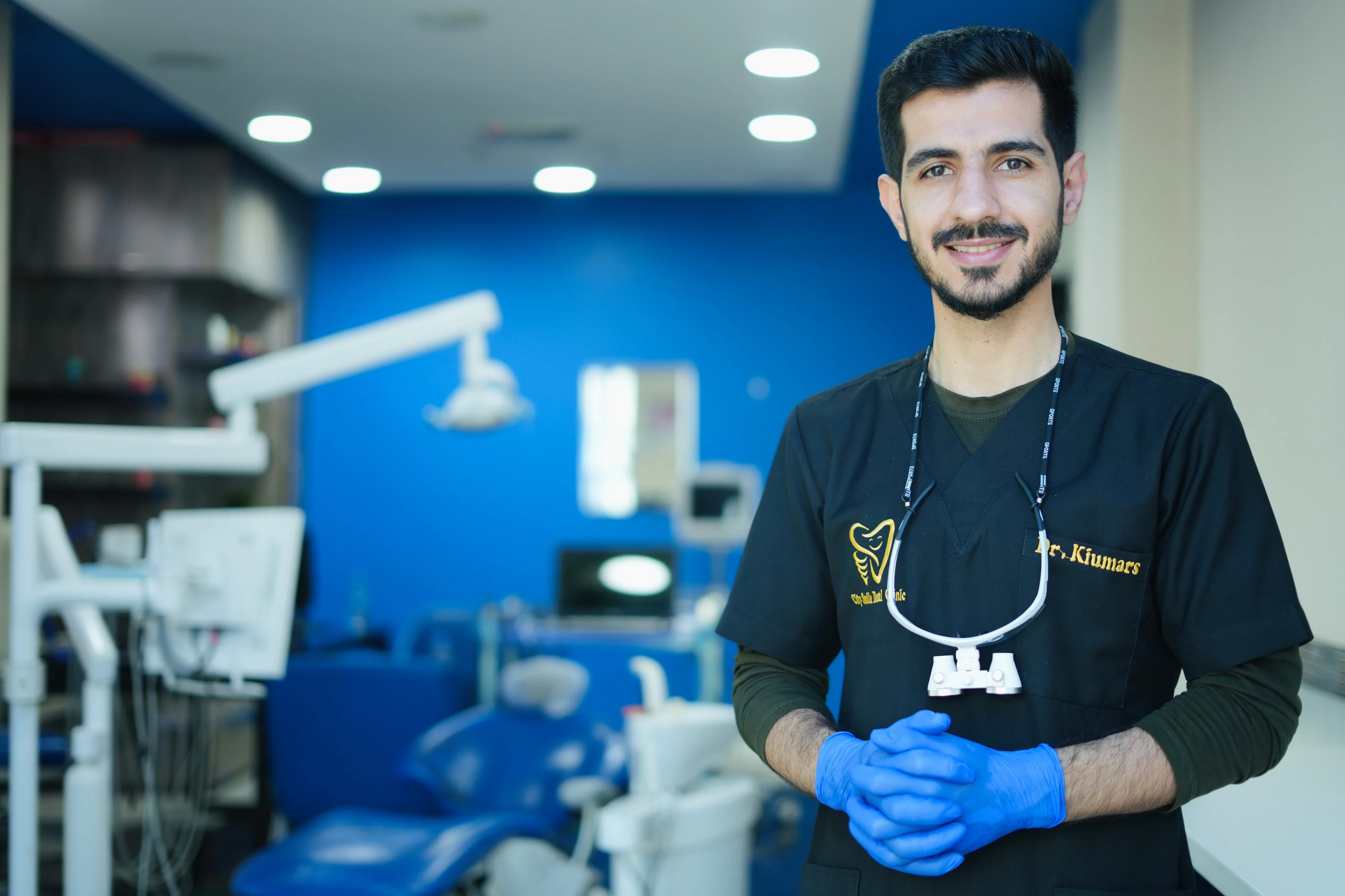 Dr. Arash Kiumars smiling in a treatment room at City Smile Dental Clinic, wearing gloves and dental loupes